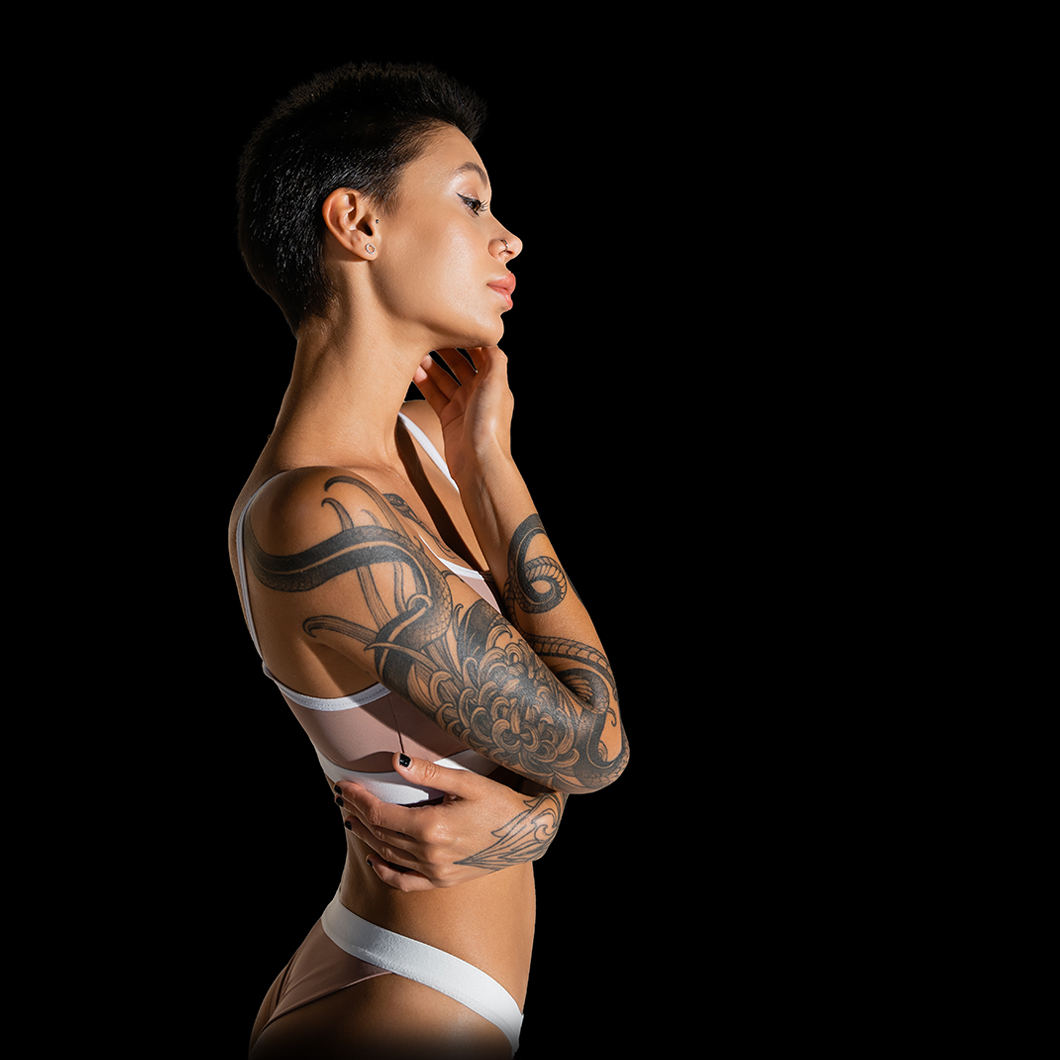 side view of tattooed woman with short brunette hair touching face while standing in bra on grey